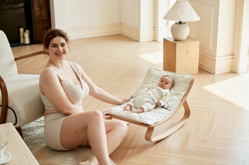 Mother in a cotton nursing bra sitting beside her newborn in a baby bouncer